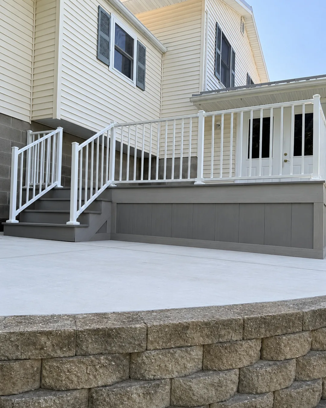 Composite deck with white aluminum railing and stairs above a concrete patio and stone retaining wall
