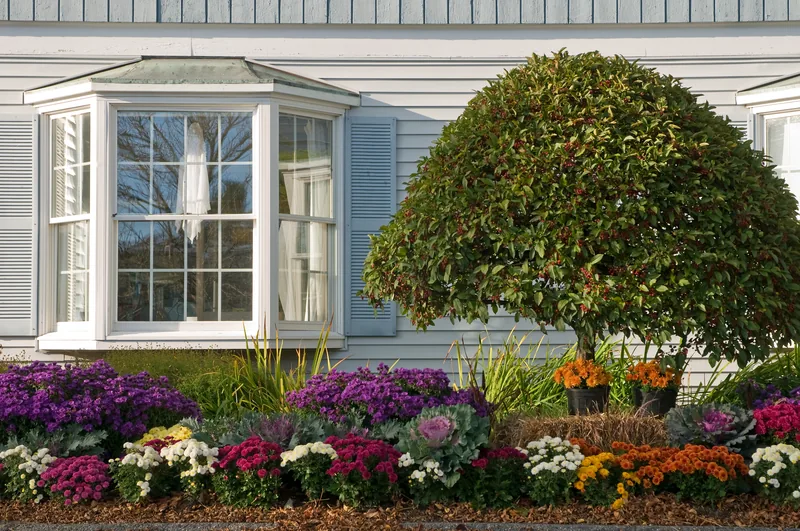 White home with a bay window, trimmed shrub and a bed of mixed flowers