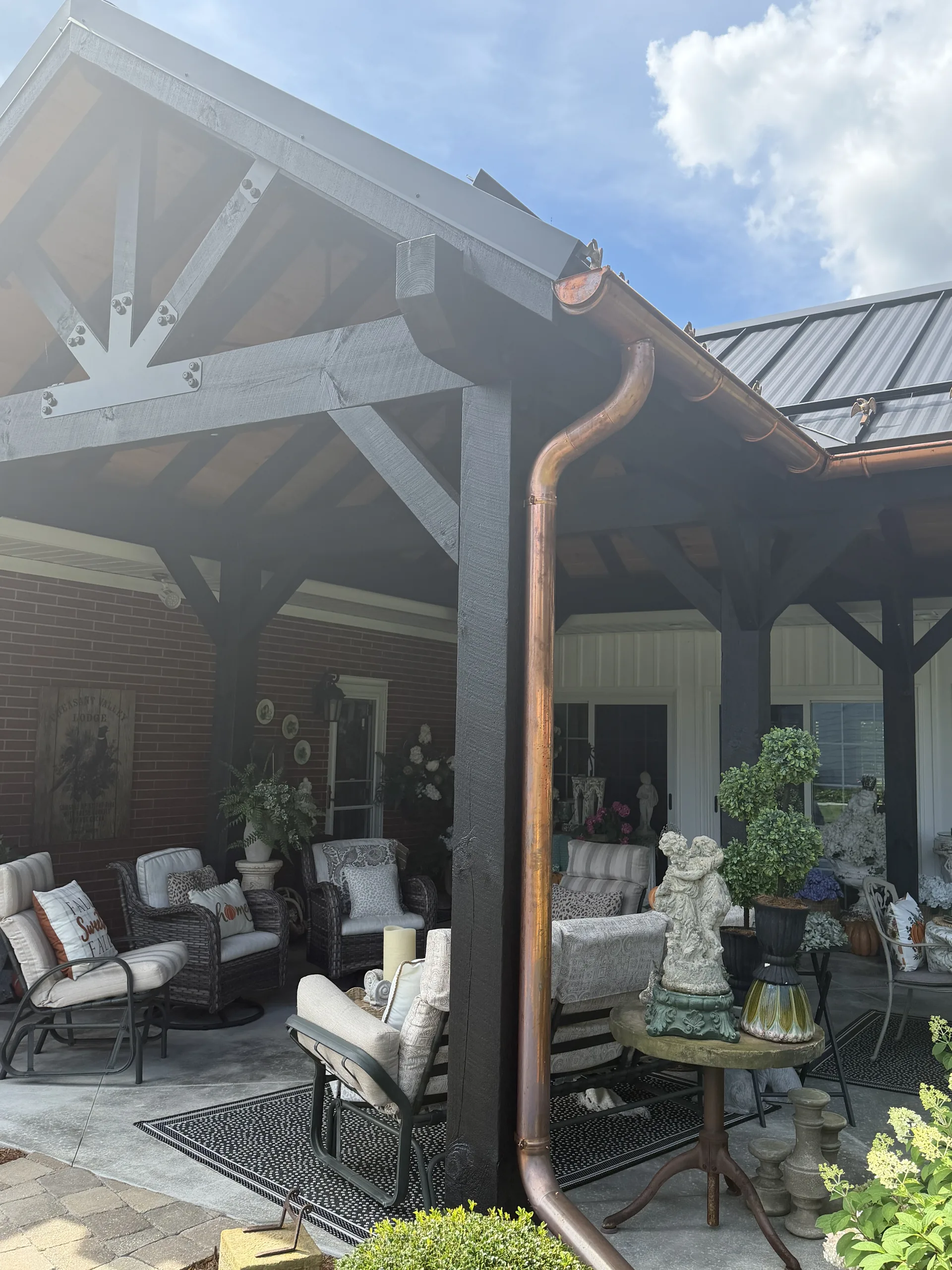 Covered patio under a timber-frame pavilion with a copper downspout, wicker seating and potted plants