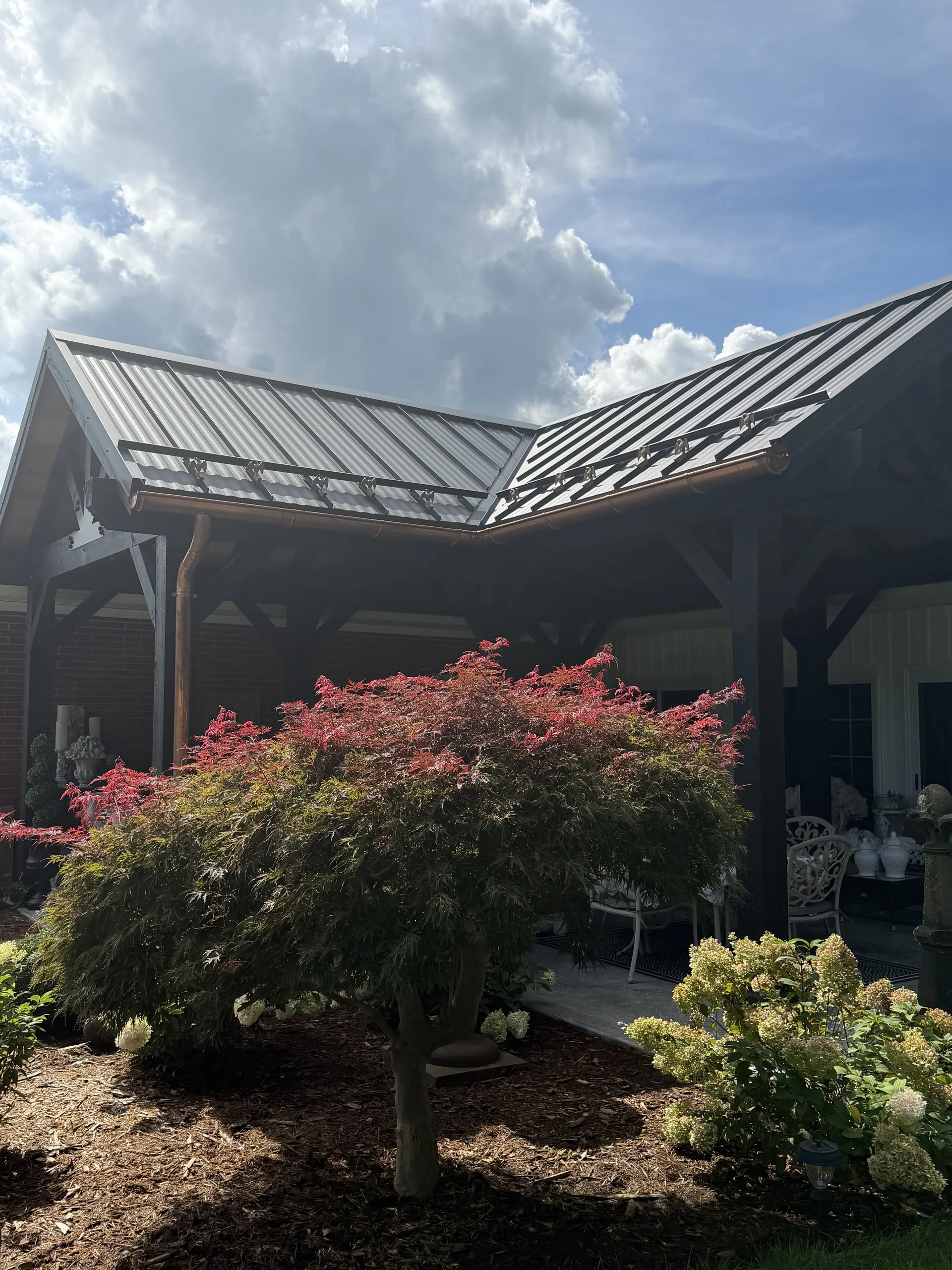 L-shaped pavilion with a black metal roof beside a brick home, with a red Japanese maple in front