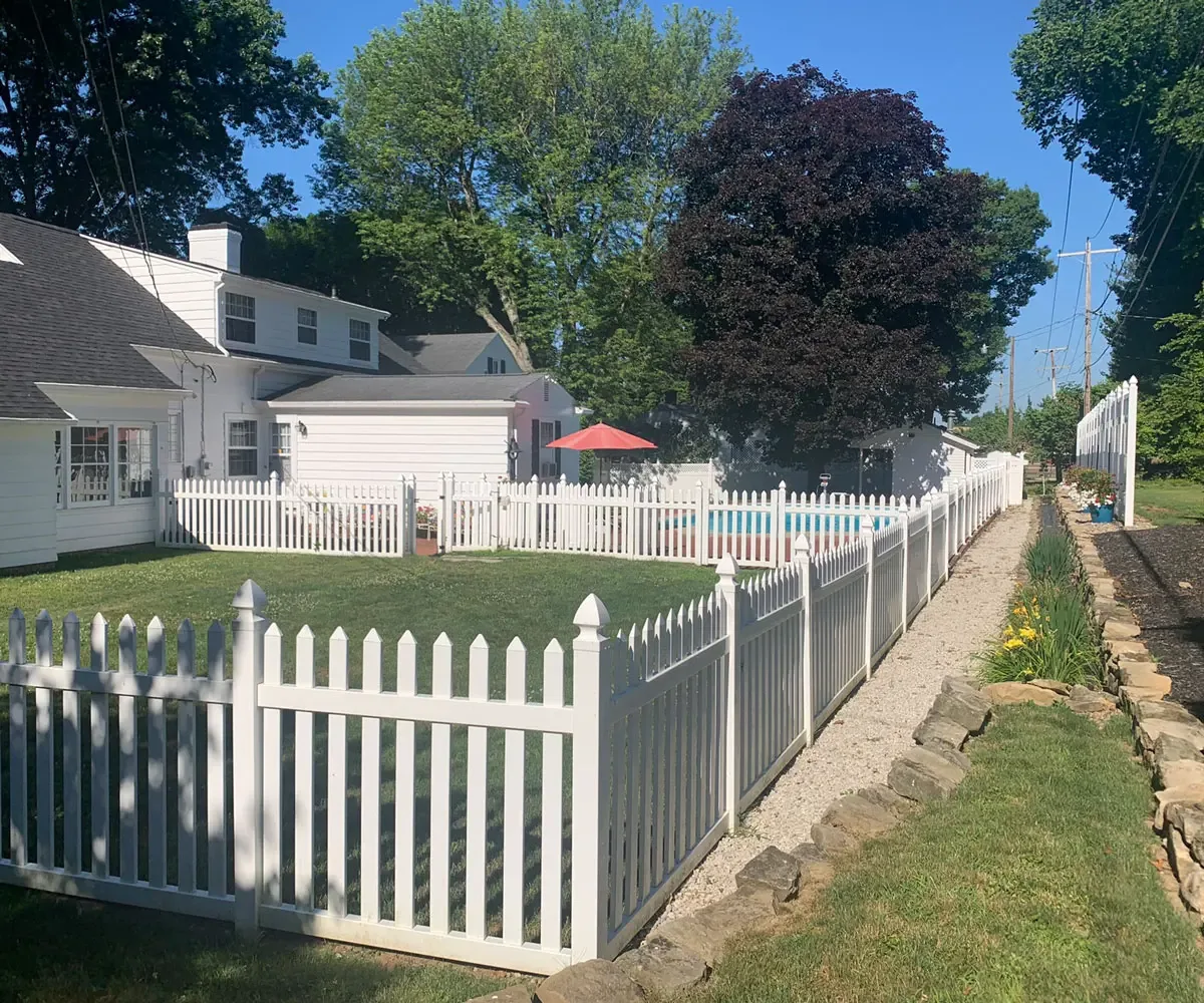 White vinyl picket fence beside a driveway with a pool and red maple