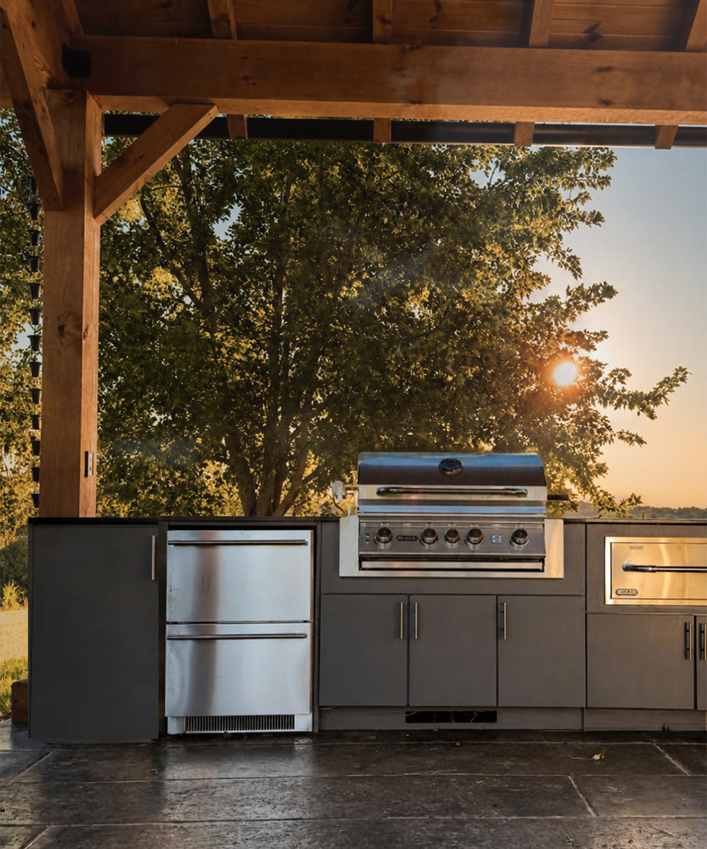 Outdoor kitchen with a stainless steel grill, refrigerator and gray cabinets beneath a timber pavilion